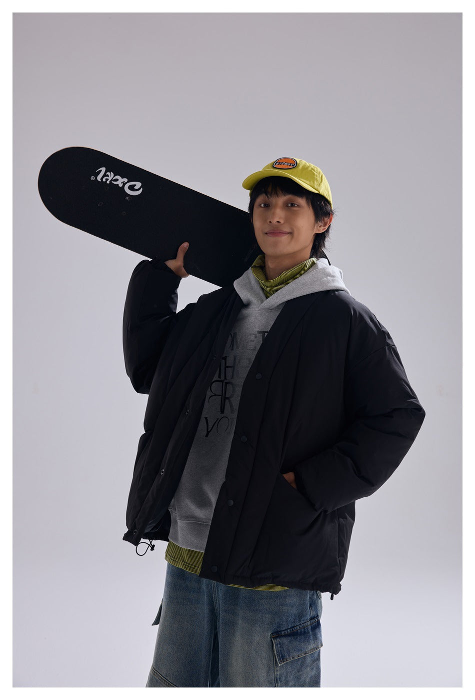 Model wearing a black padded jacket over a gray hoodie, holding a skateboard on his shoulder.