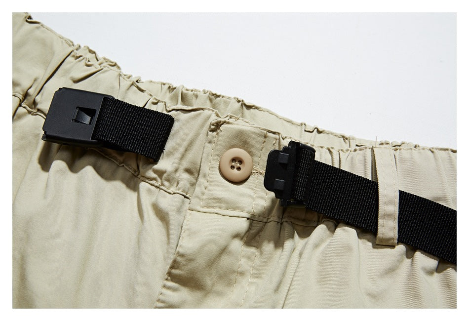 Close-up of beige trousers showing an elastic waistband, black webbing belt and buckle, and a button.