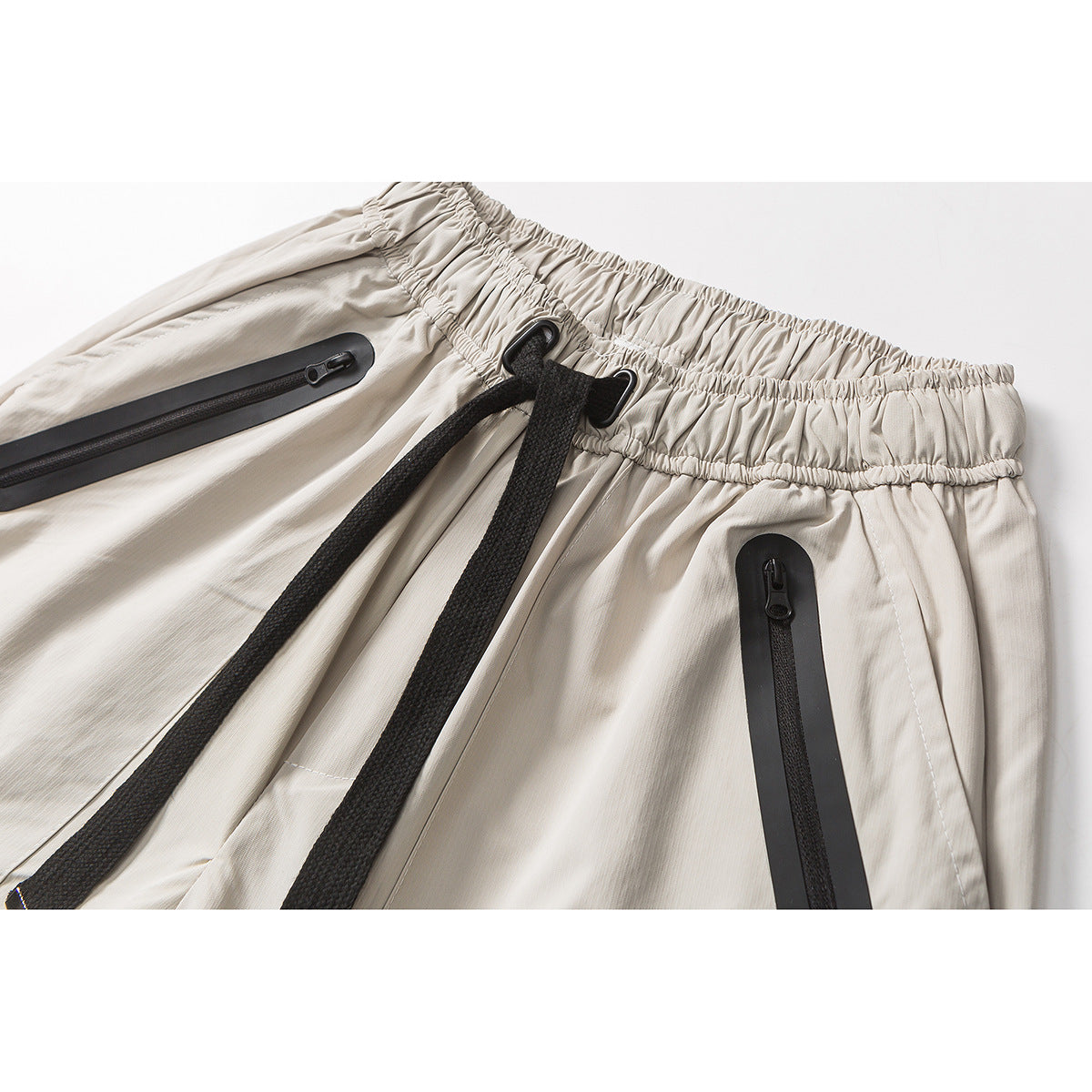 Close-up of light beige shorts with a gathered waistband, black drawcord, and black-trimmed zip pockets.