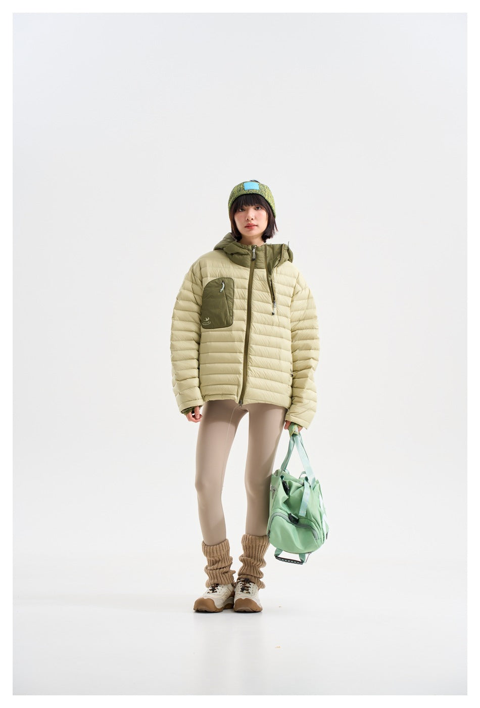 Model wearing a pale yellow-green hooded puffer jacket with an olive chest pocket, shown standing with a mint bag.