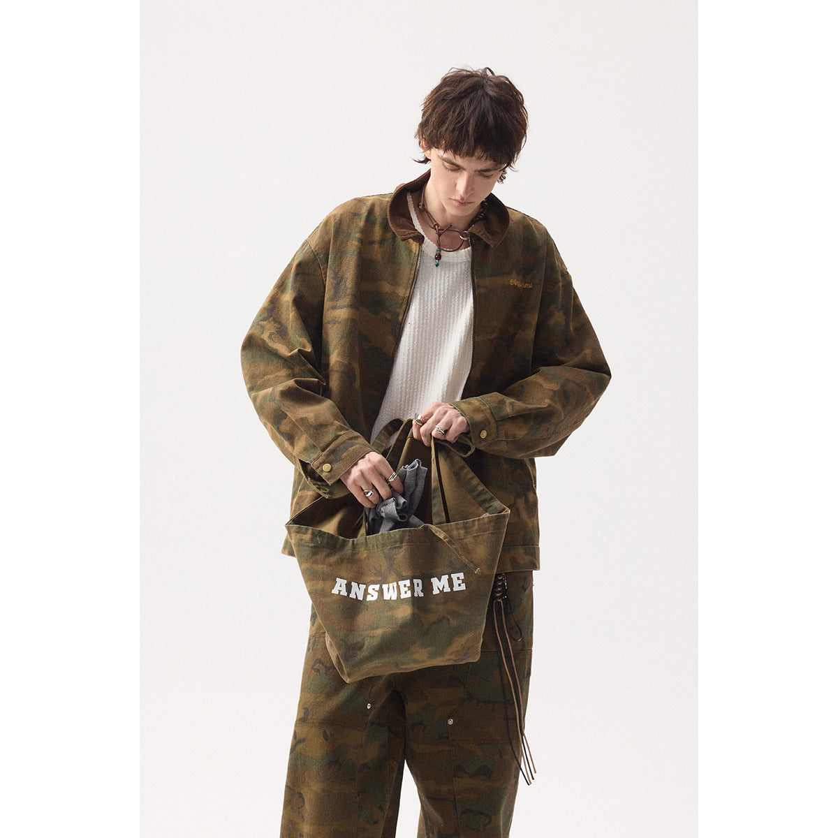 Model wearing a brown-green camouflage zip jacket and loose matching trousers, carrying a matching tote printed “ANSWER ME.”