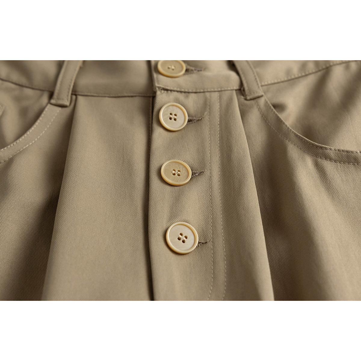 Close-up of taupe trousers with four tan buttons along the front fly and curved front pocket edges.