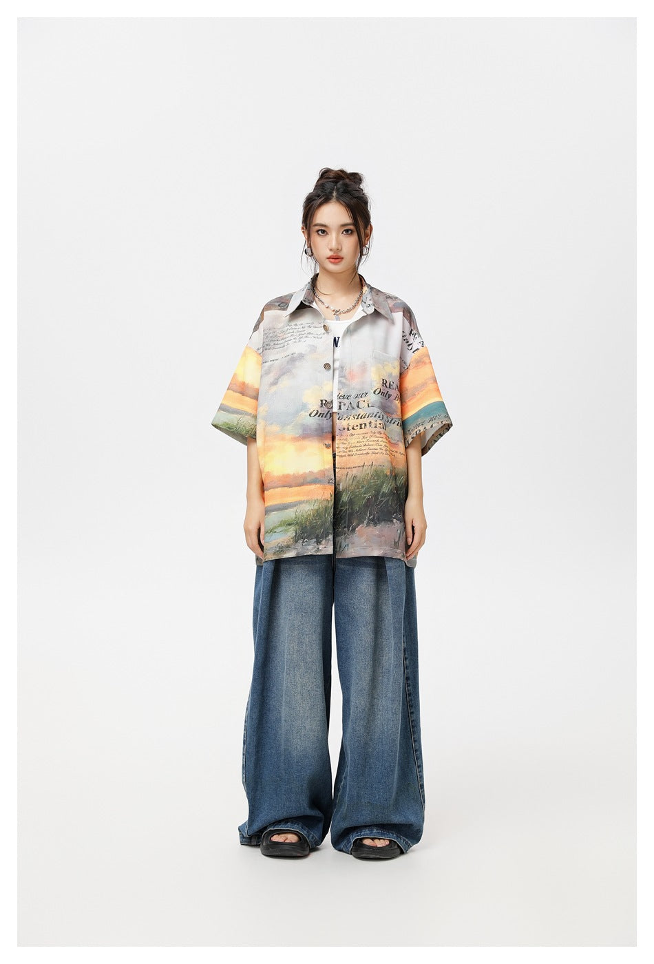 Model wearing a sunset landscape print short-sleeve shirt open over a white top with wide-leg blue jeans, front view