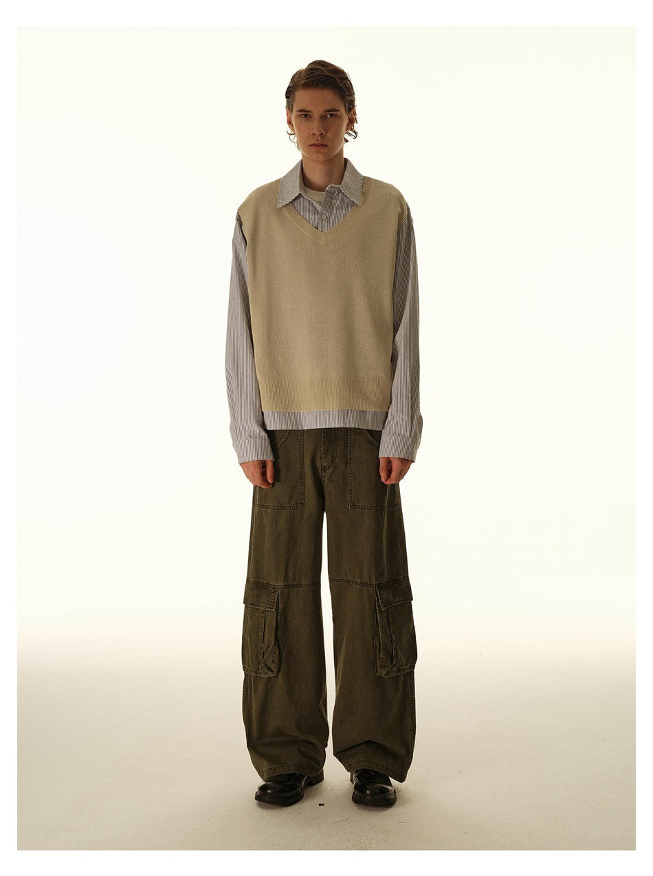 Front view of a model in a tan V-neck sweater vest over a gray striped shirt with loose dark olive cargo pants.