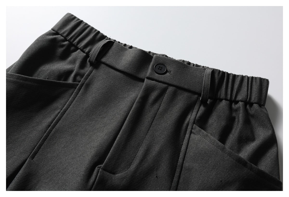 Close-up of dark trousers showing a buttoned waistband, belt loops, pleats and front pockets.