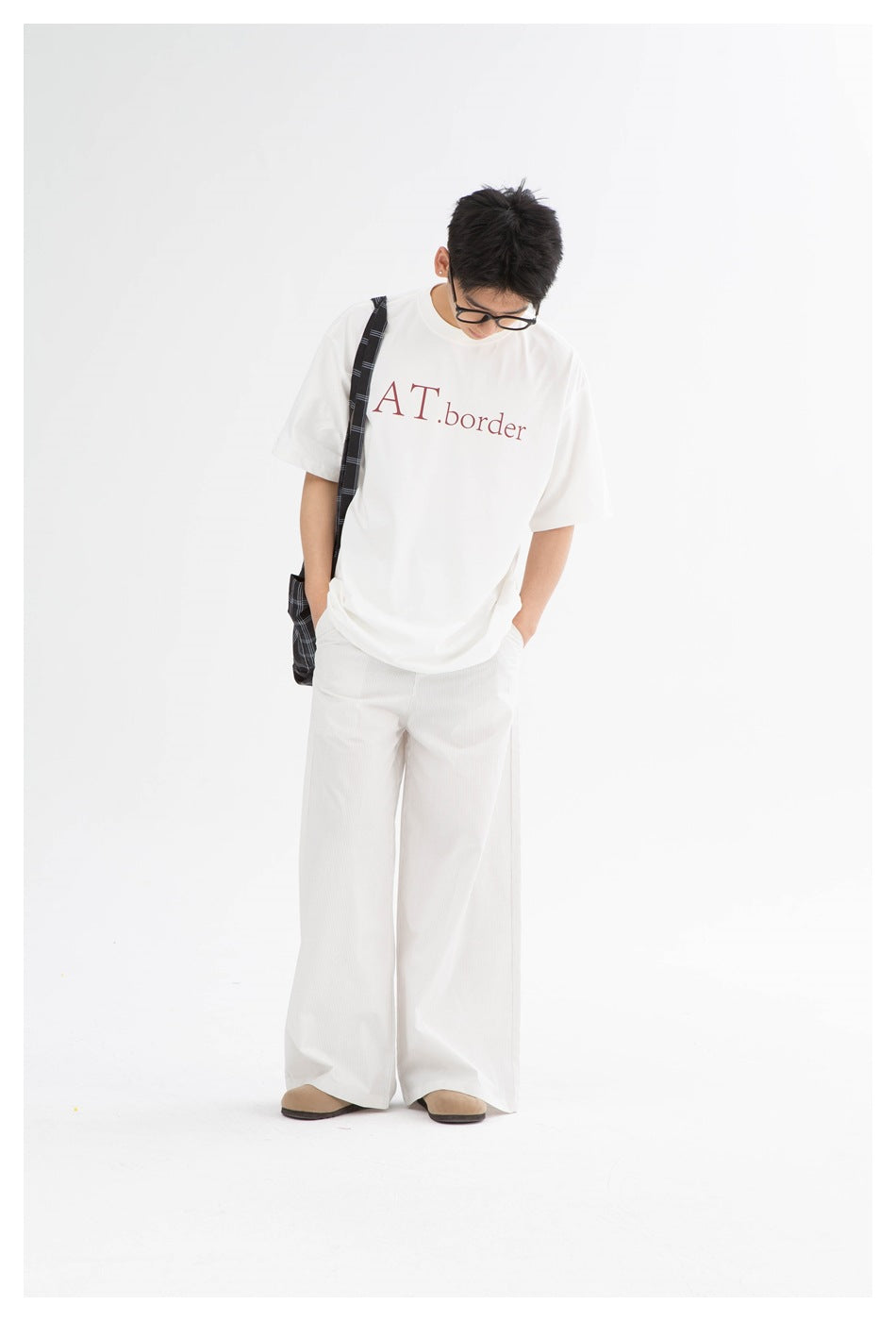 Full-length view of a model in a white oversized graphic T-shirt, wide-leg white pants, and a black plaid shoulder bag.