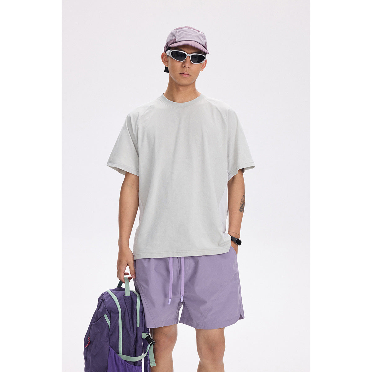 Model wearing a loose light gray T-shirt and lavender shorts, with a cap, sunglasses, and backpack.