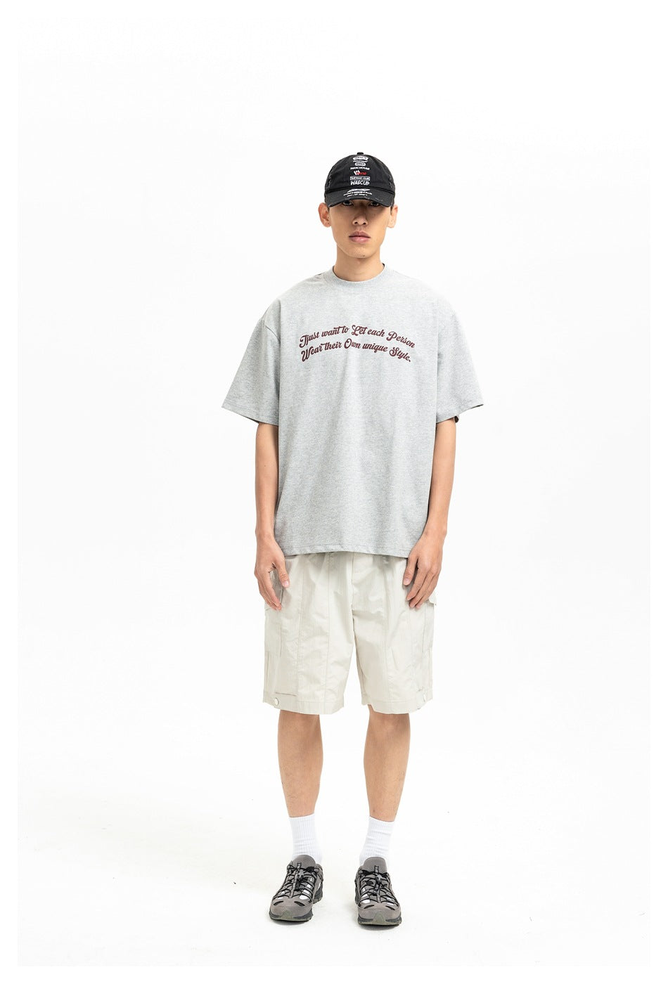 Front view of a model in a light gray T-shirt with burgundy script, cream cargo shorts, and a black cap.