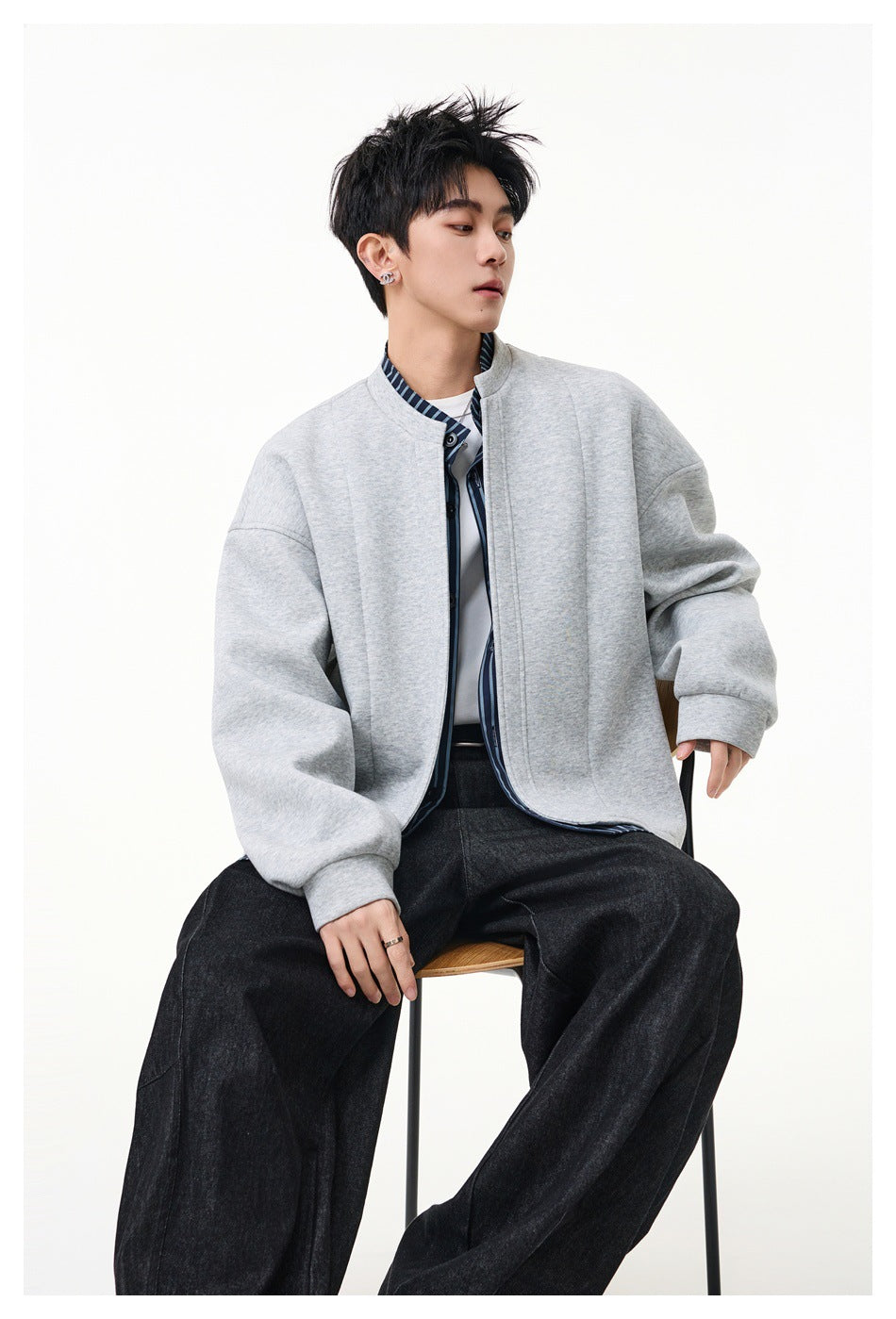 Seated model wearing an open light gray jacket with striped trim, a shirt, and wide black trousers.