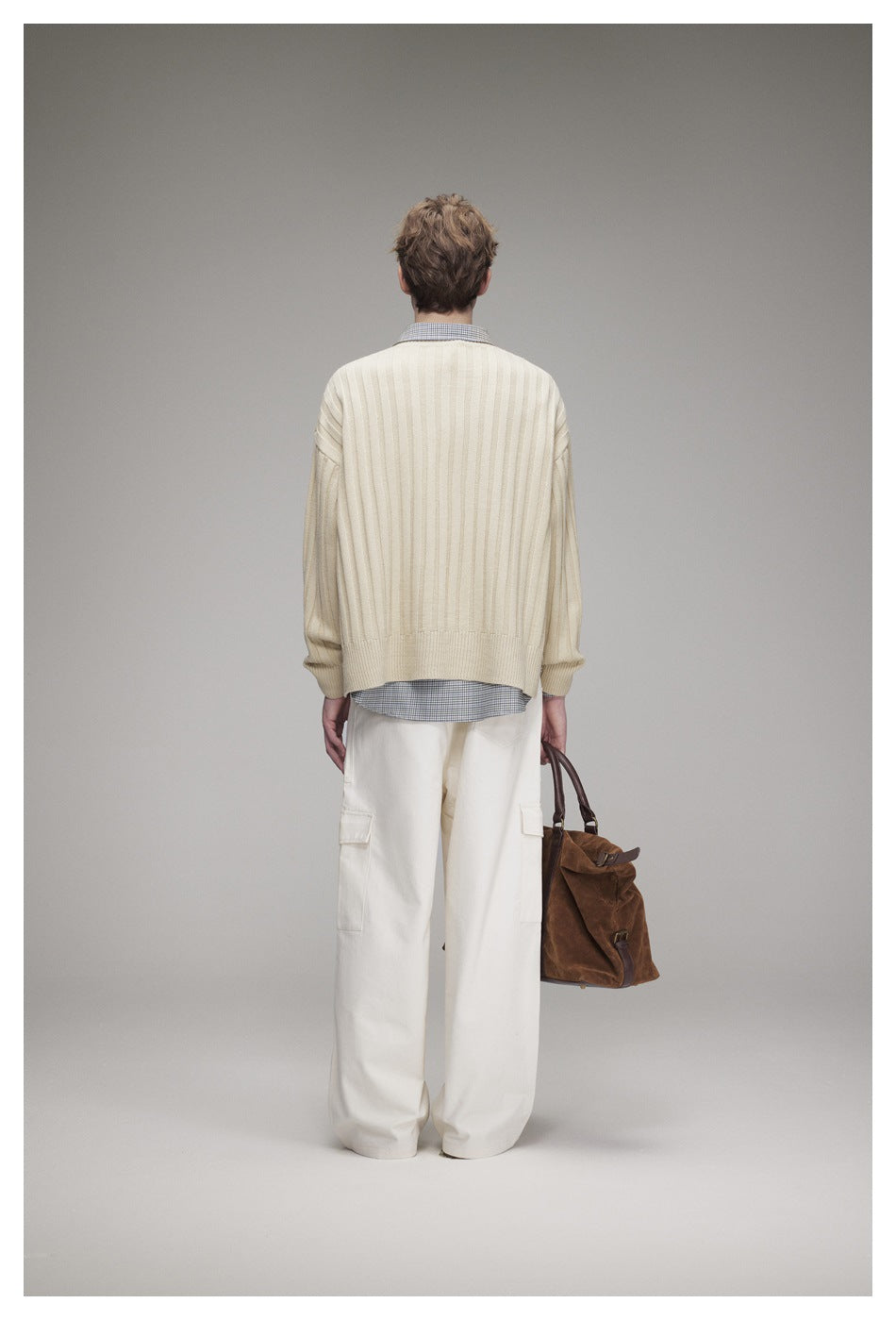 Rear view of a model wearing a cream ribbed sweater over a checked shirt and loose white cargo trousers.