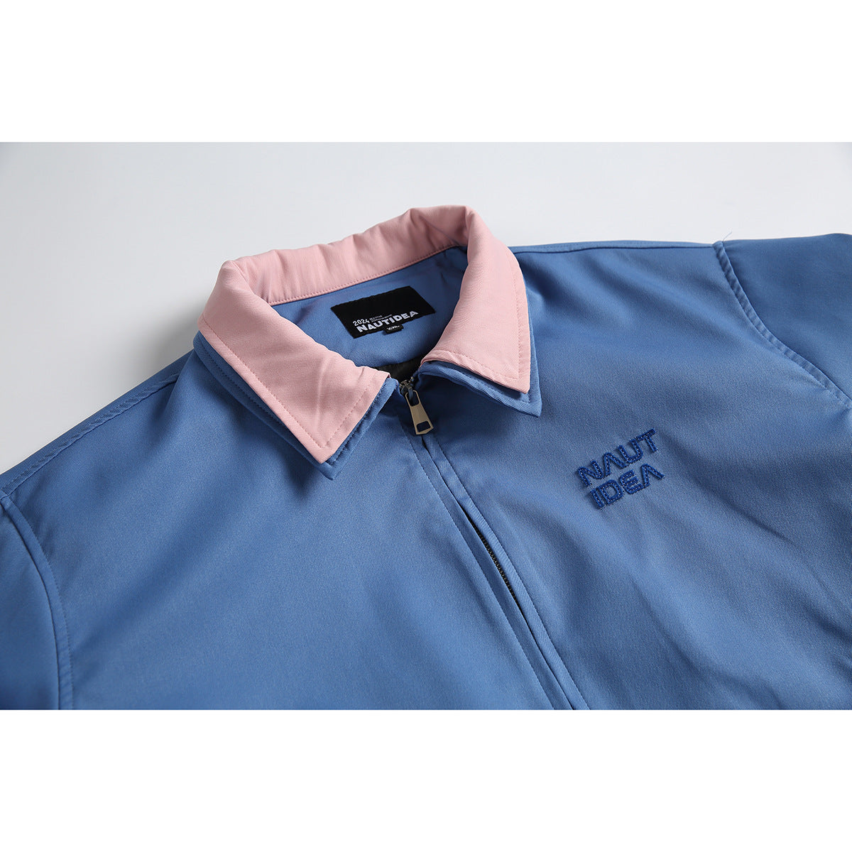 Close-up of a blue zip jacket with a pink collar, chest embroidery, and a visible neck label.