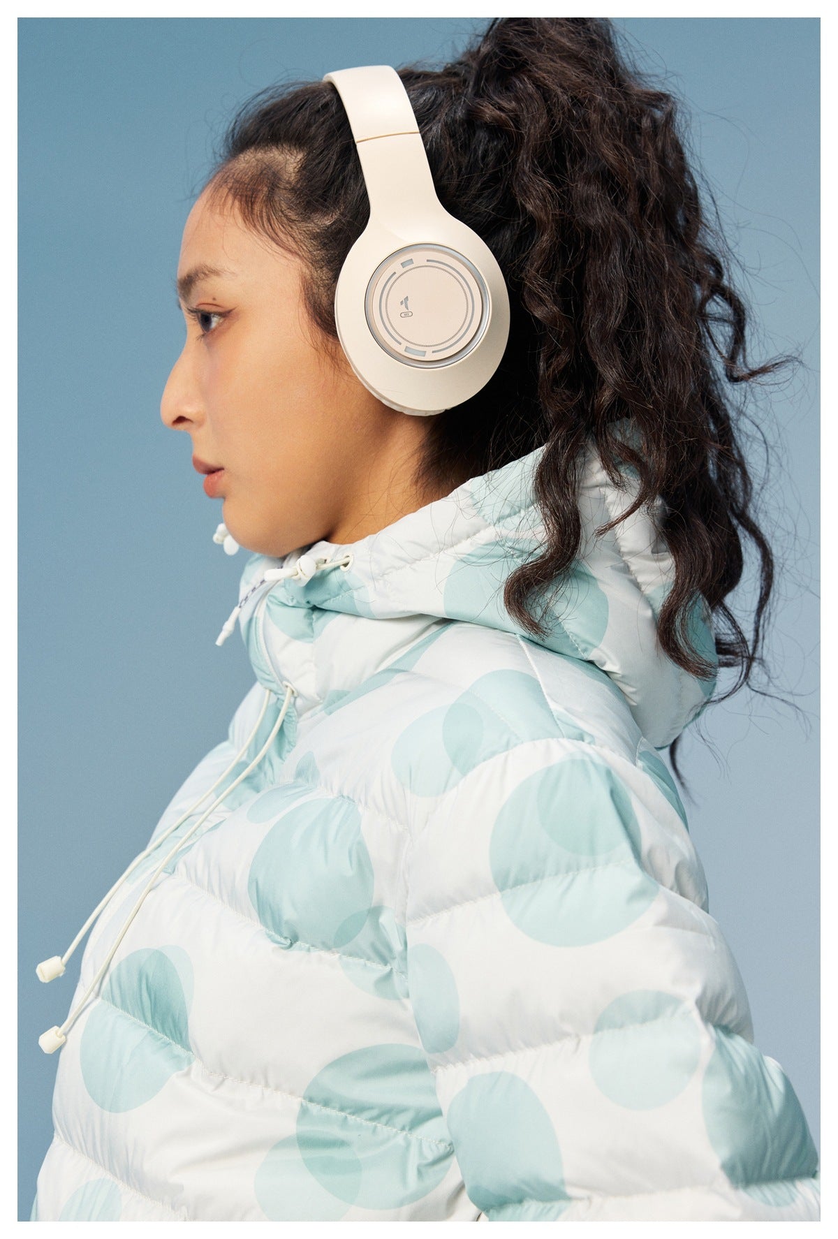 Close side view of a model wearing a white puffer jacket with pale aqua circles and a high drawcord hood.