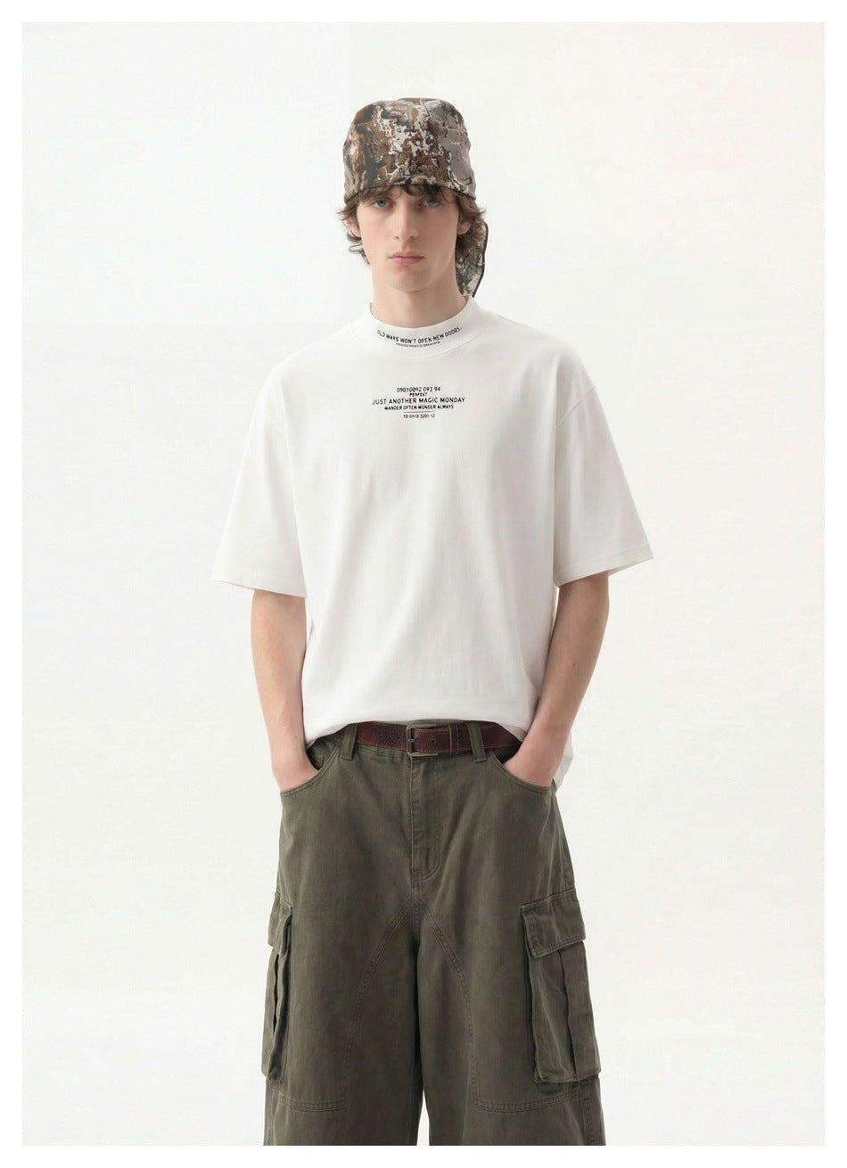 Model wearing a white oversized T-shirt with small black chest and collar text, olive cargo pants, and a camouflage