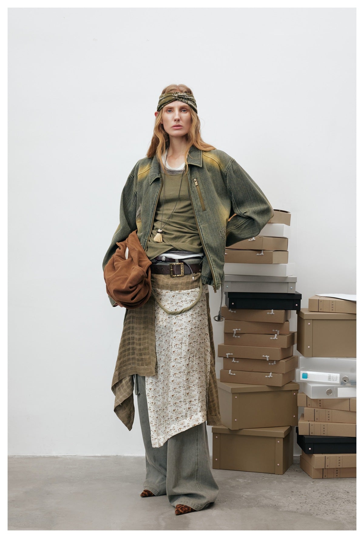 Model wearing the washed green denim zip jacket open over a olive top with layered skirt, wide trousers and boxes behind