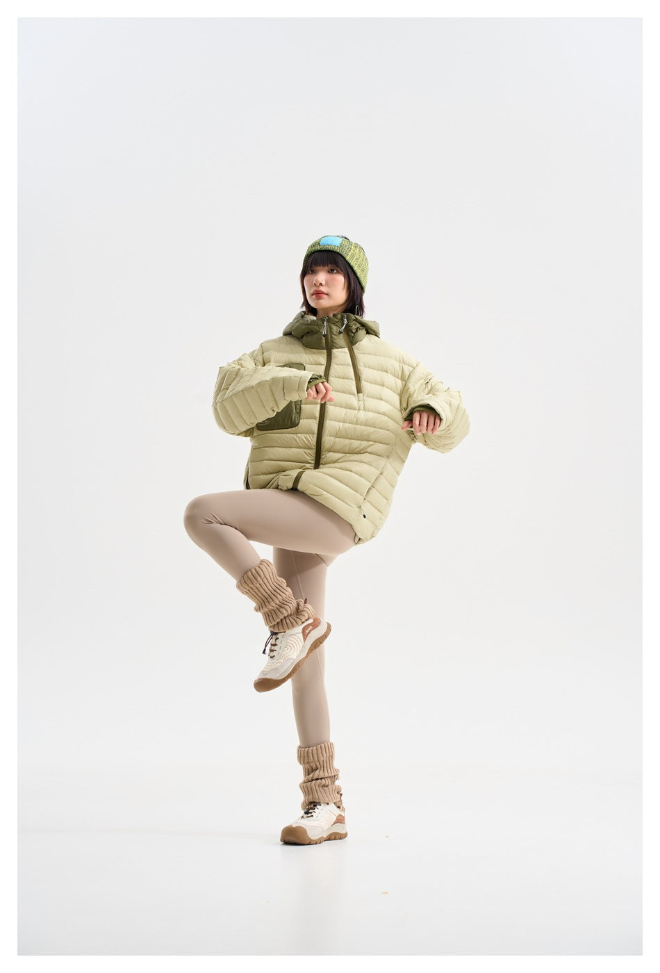 Model wearing a pale yellow-green hooded puffer jacket with an olive chest pocket, posing with one knee raised.
