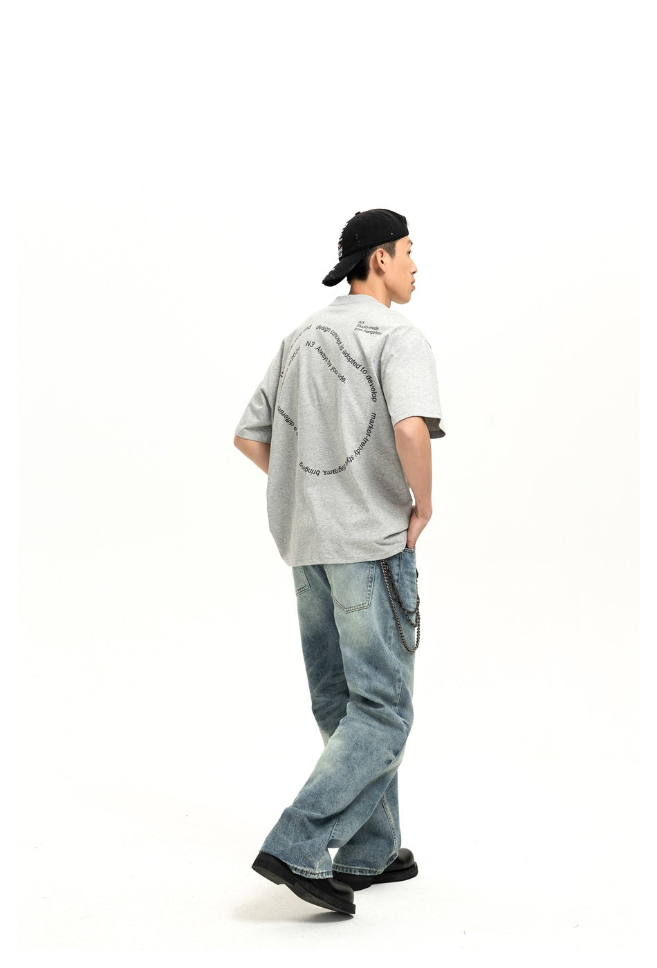 Full-length side-back view of a model wearing a gray T-shirt with black circular back text and loose blue jeans.