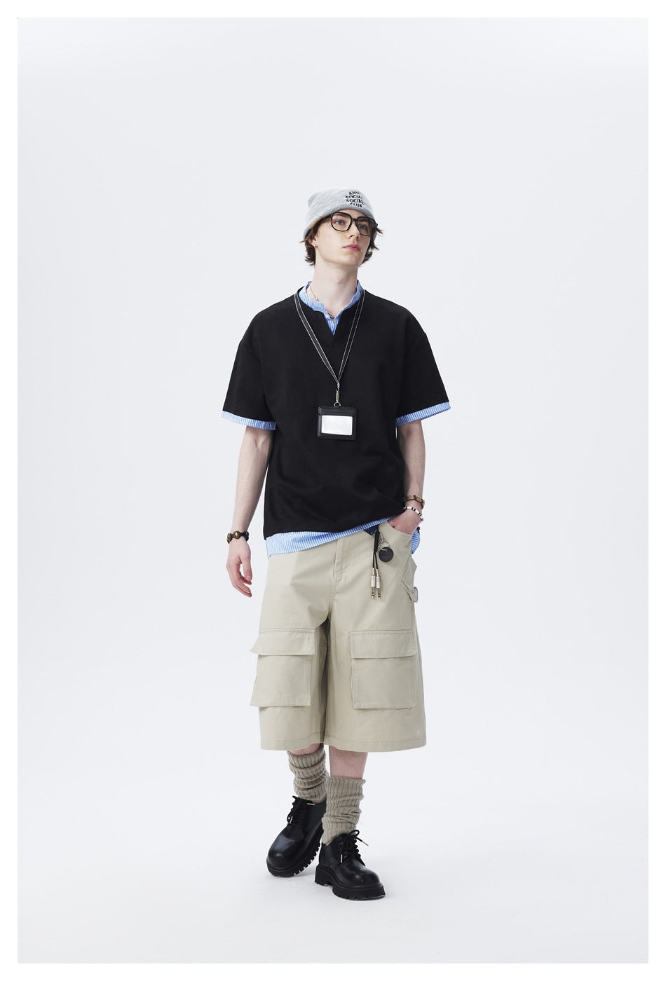Man wearing black layered striped-trim tee with beige cargo shorts, beanie, glasses and lanyard, full-length front view