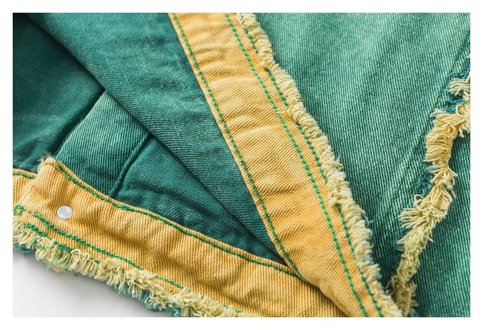 Close-up of teal denim with mustard-yellow contrast panels, green stitching, frayed edges and a small metal snap.