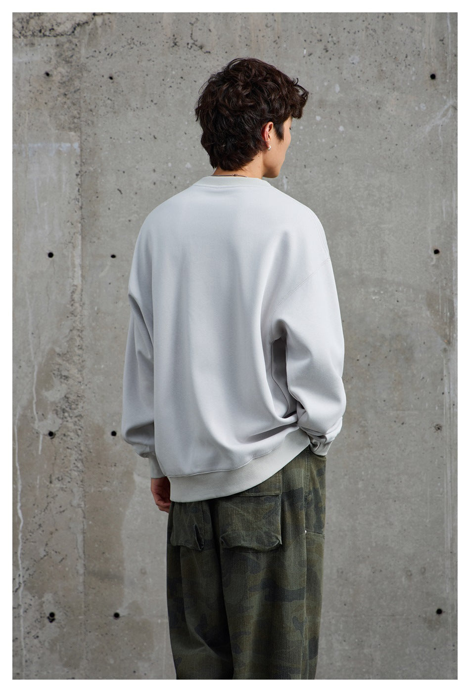 Back view of a model wearing a plain light gray sweatshirt and loose dark camouflage pants.