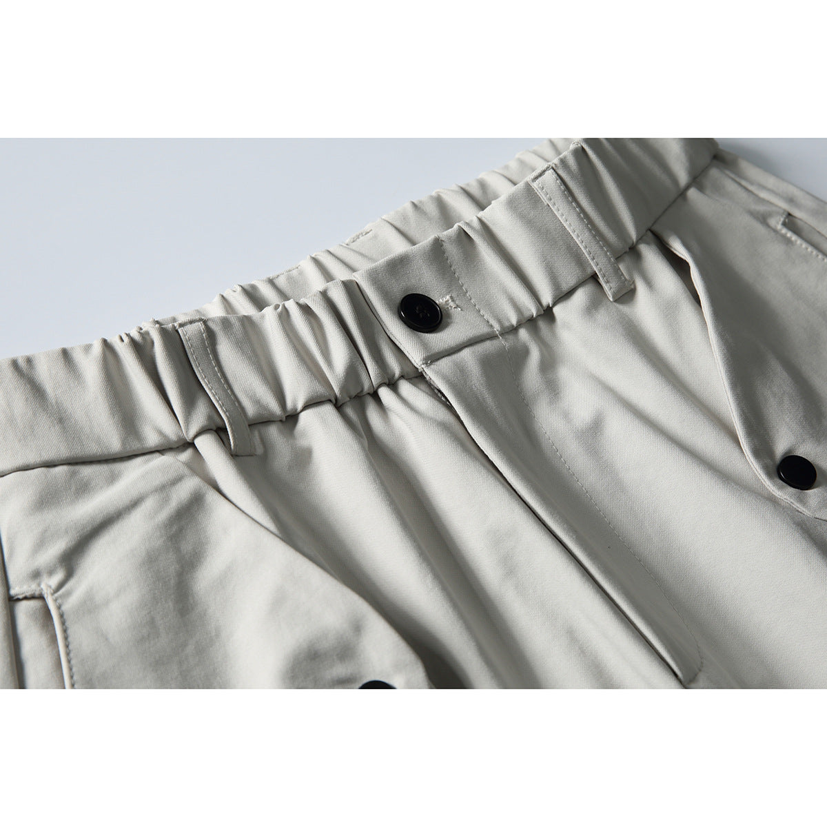 Close-up of light gray trousers with a gathered waistband, belt loops, pleats, and black buttons.
