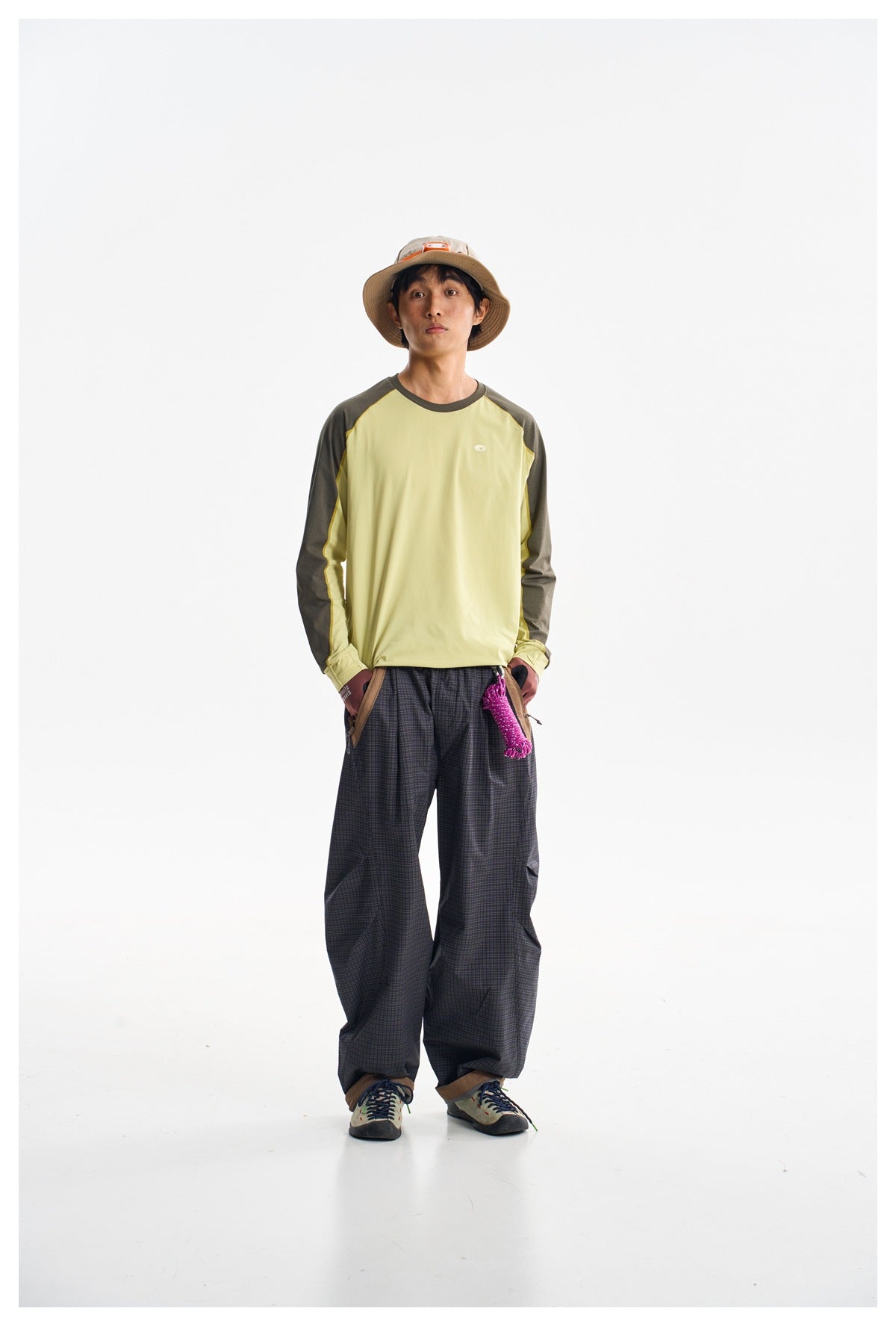 A man models a pale yellow long-sleeve raglan shirt with dark olive sleeves and loose dark plaid pants.