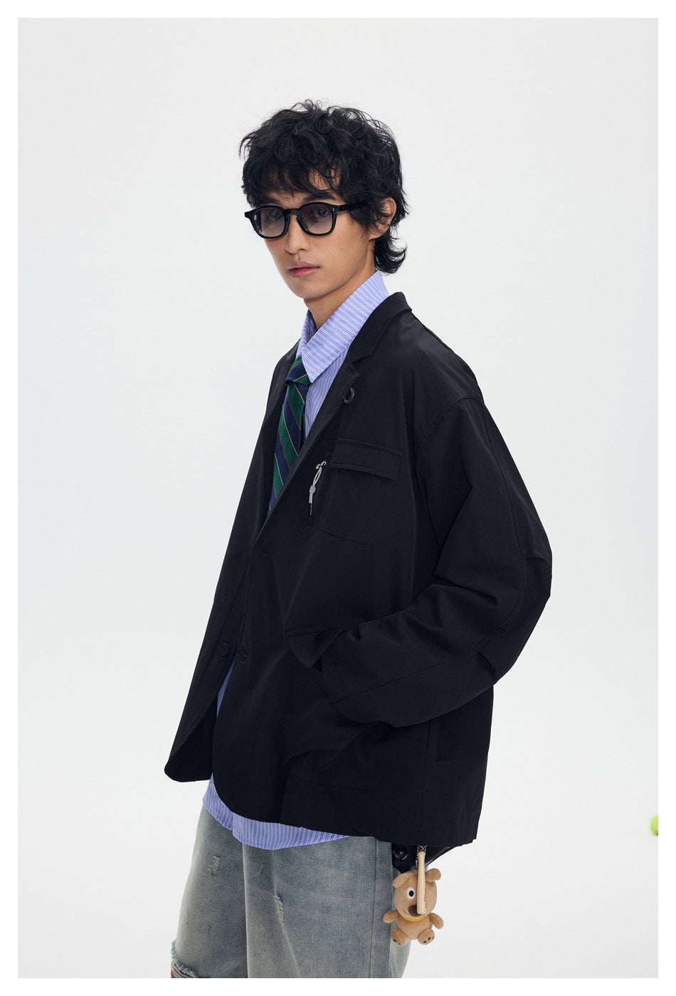 Model wearing a black oversized blazer with a zip chest pocket, striped shirt, tie, and distressed jeans.