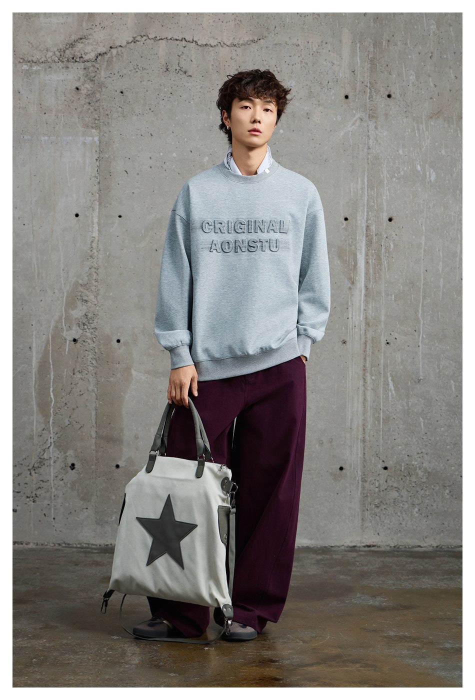 Model wearing a light gray crewneck sweatshirt with raised chest lettering and dark plum trousers.