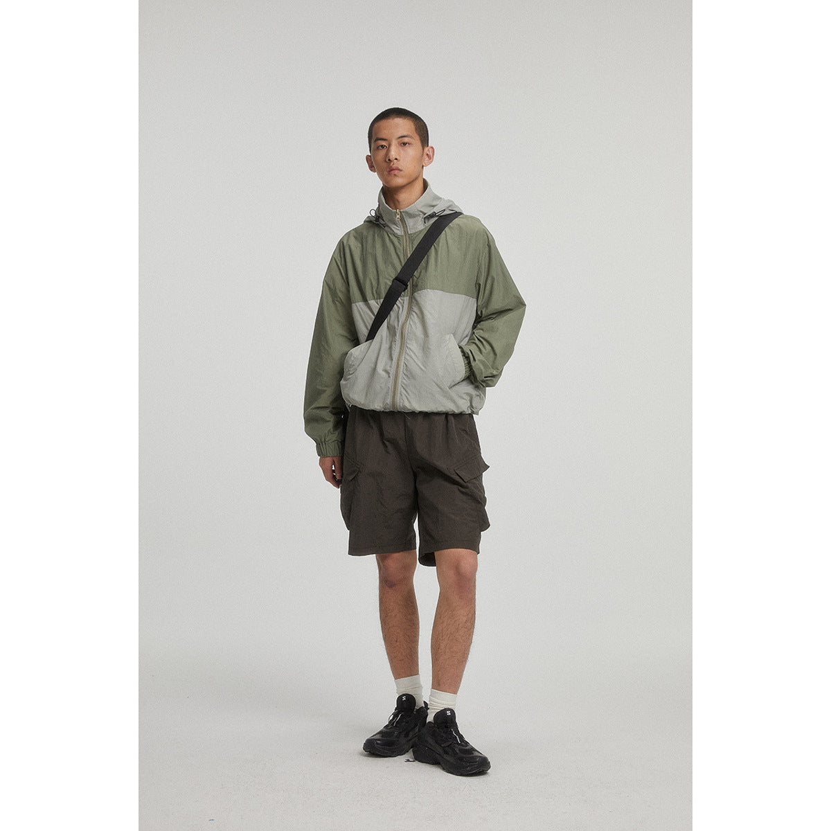 Model wearing an olive green and light gray hooded color-block jacket with dark cargo shorts.