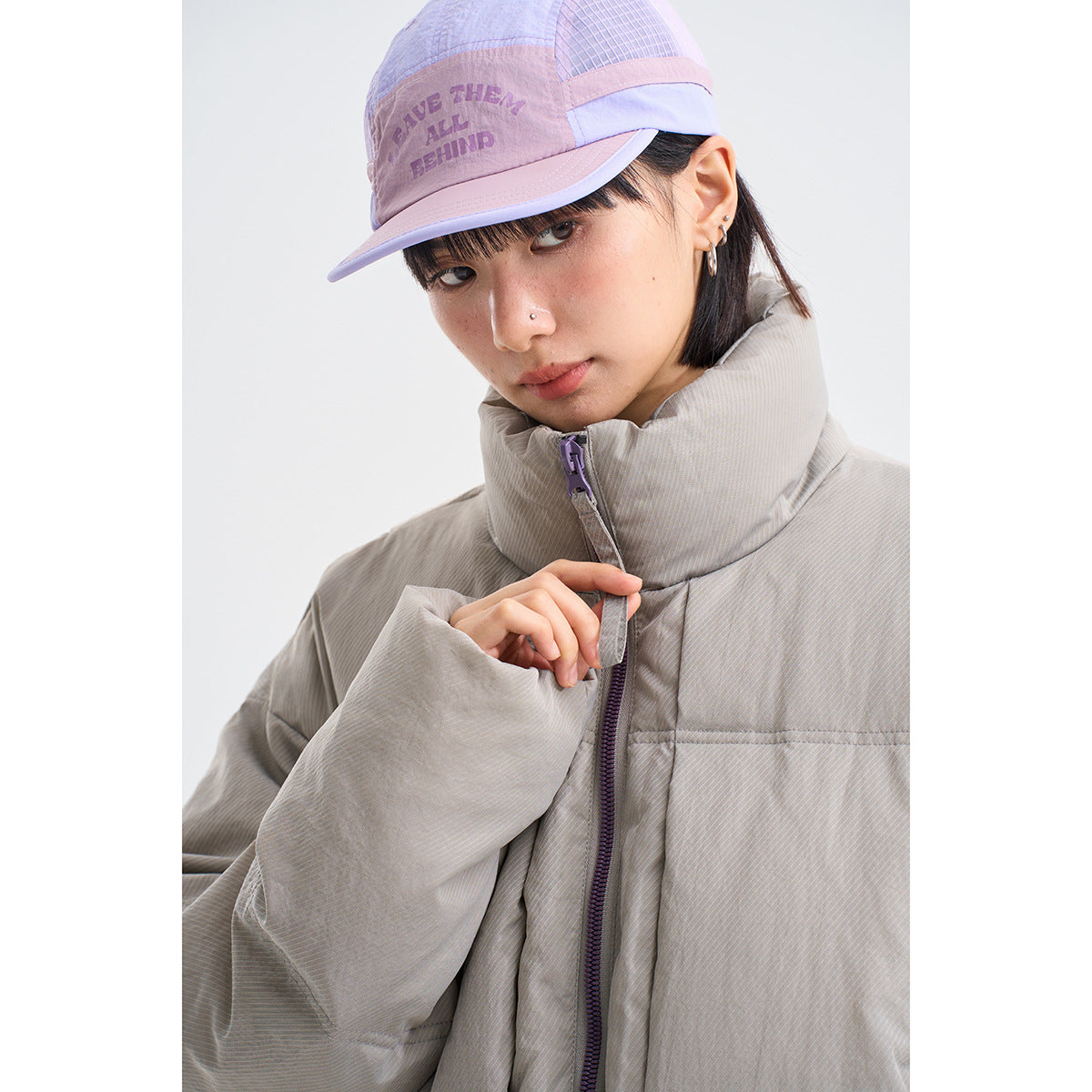 Close-up of a model in a light gray high-collar puffer jacket and lavender cap with English text.