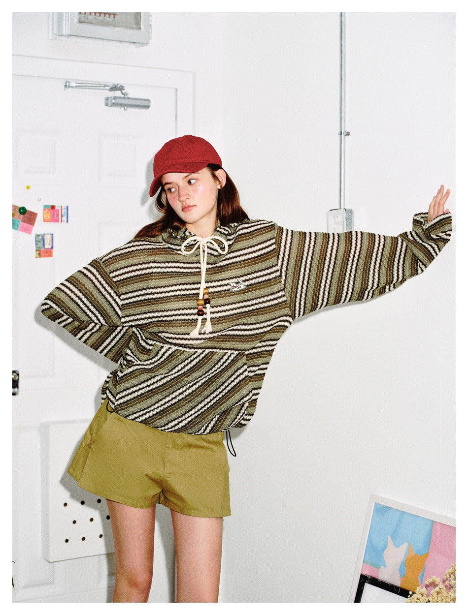 Model in an olive, cream and black striped hooded pullover and olive shorts, posing with one arm outstretched.