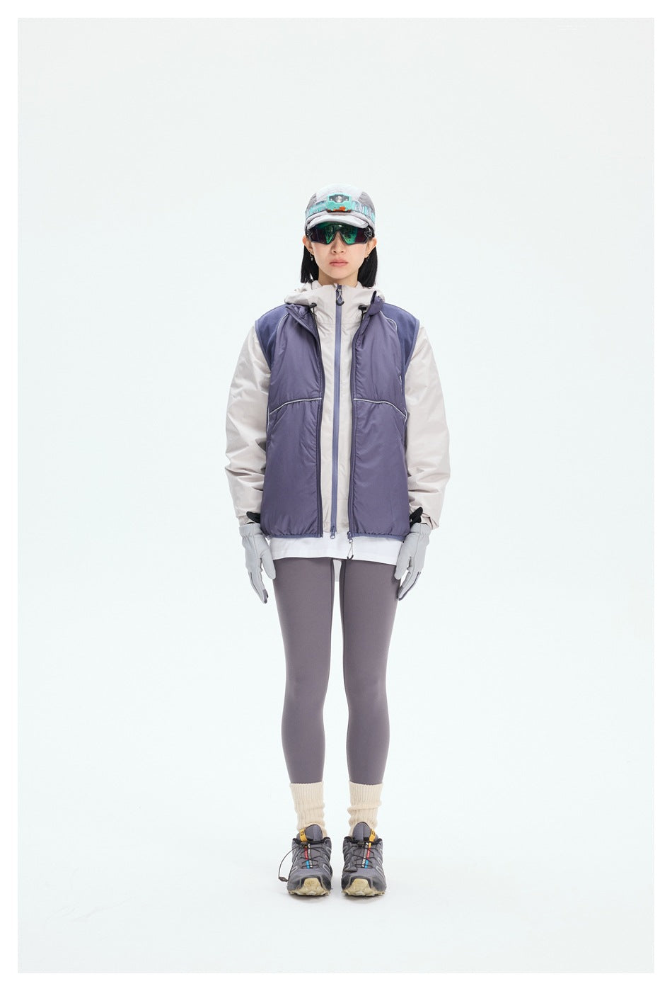 Model standing in a purple padded vest layered over a light hooded jacket with leggings and hiking shoes