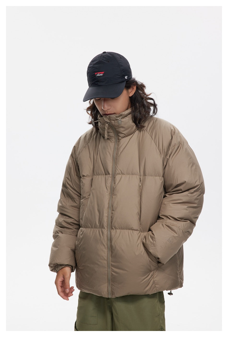 Model wearing a taupe puffer jacket zipped to its high collar, with olive pants.