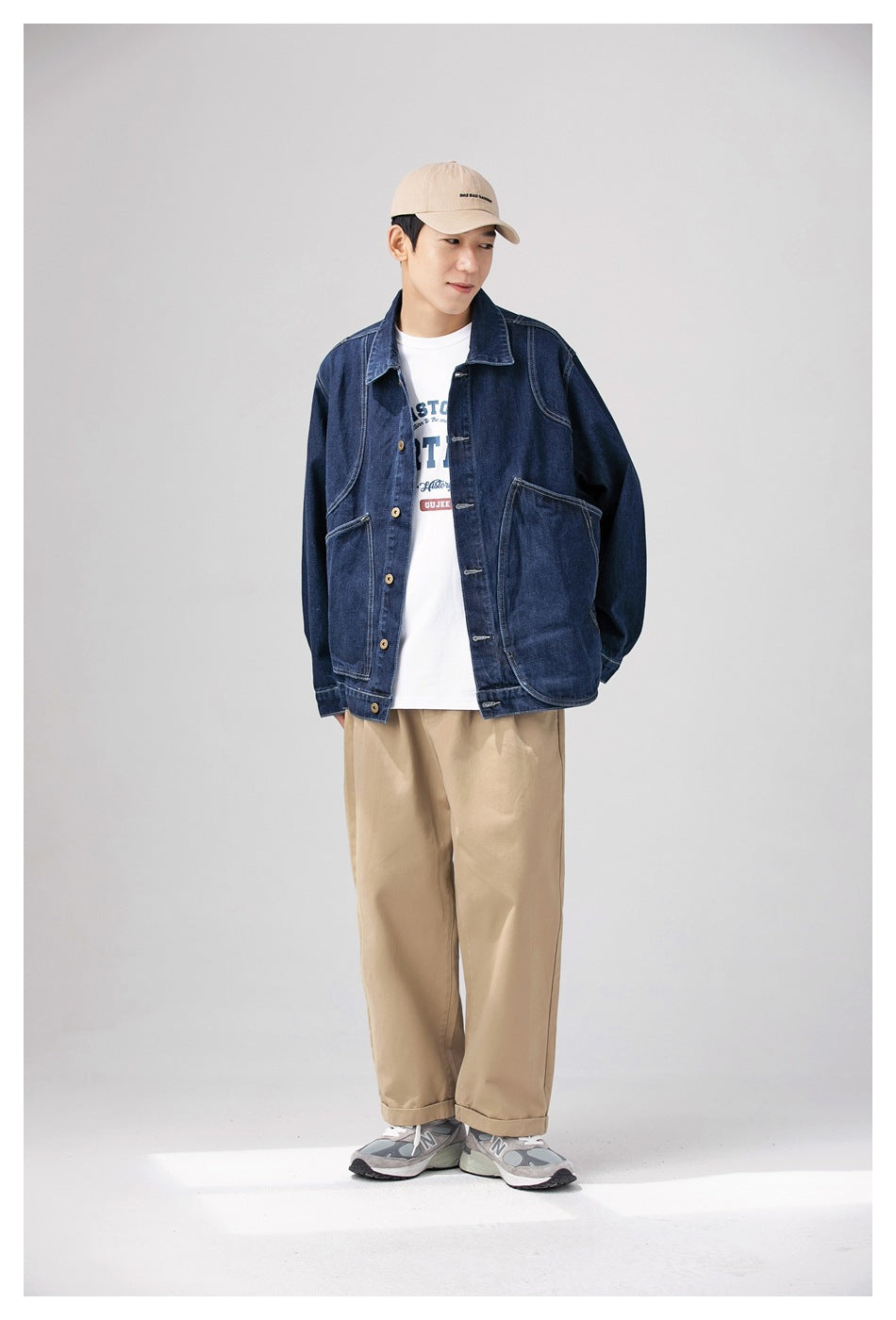 Model wearing a dark denim jacket open over a white graphic tee with khaki pants and a beige cap, three-quarter view