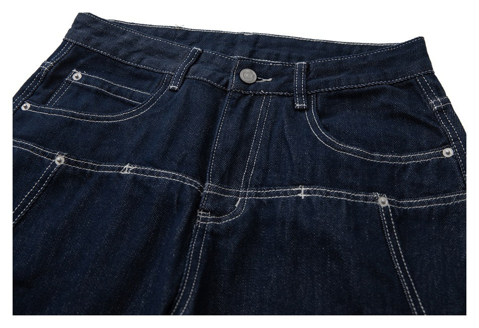 Close-up of dark indigo jeans showing the front waistband, button, riveted pockets and contrast stitching.