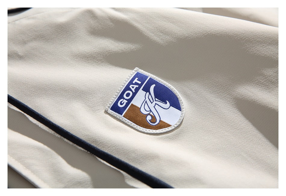 Close-up of an ivory jacket showing a blue, white, and brown embroidered GOAT patch and dark piping.