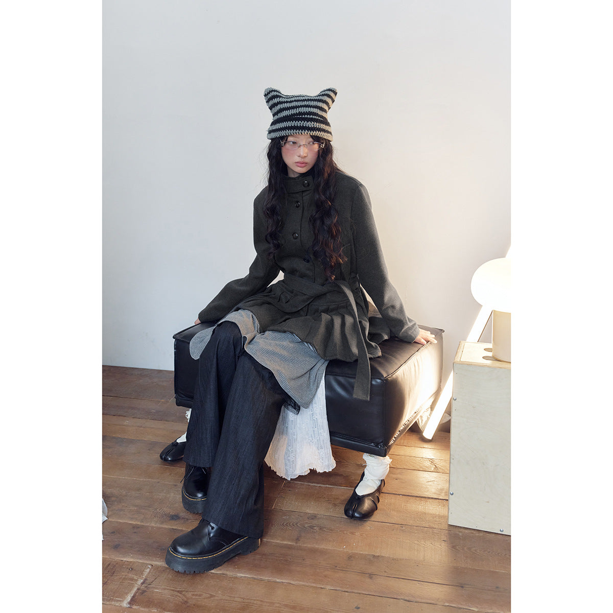 Model seated in a charcoal belted coat with a pleated hem, striped knit hat, layered skirt and wide-leg trousers.