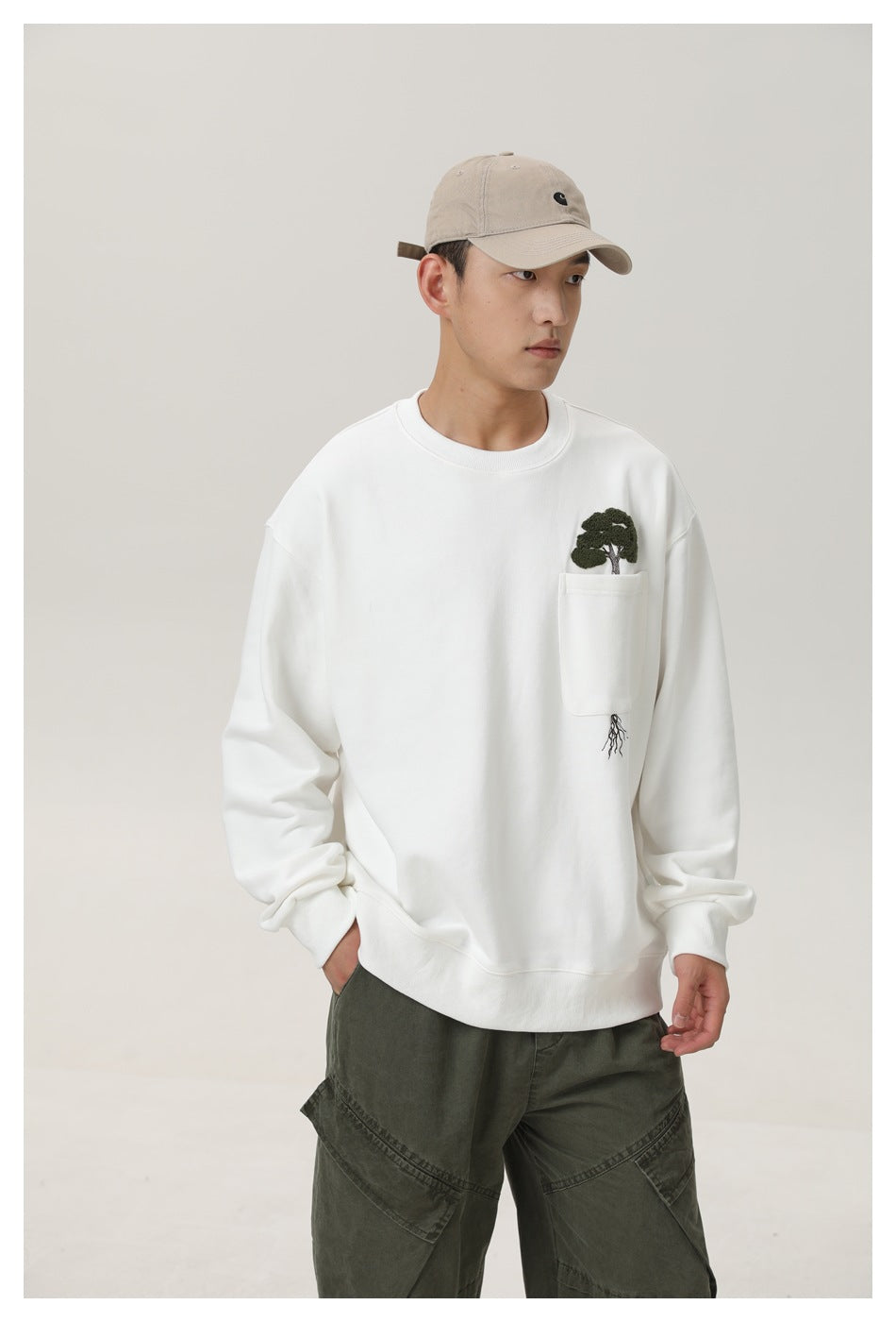 Model wearing a white crewneck sweatshirt with embroidered tree pocket, beige cap and olive cargo pants