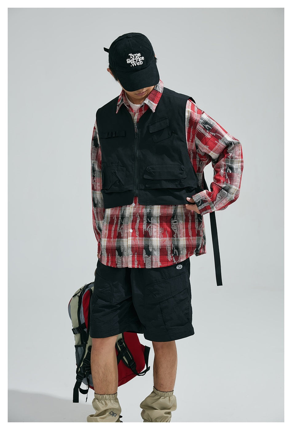 Model wearing a black utility vest over a red plaid shirt, with black shorts and a cap.