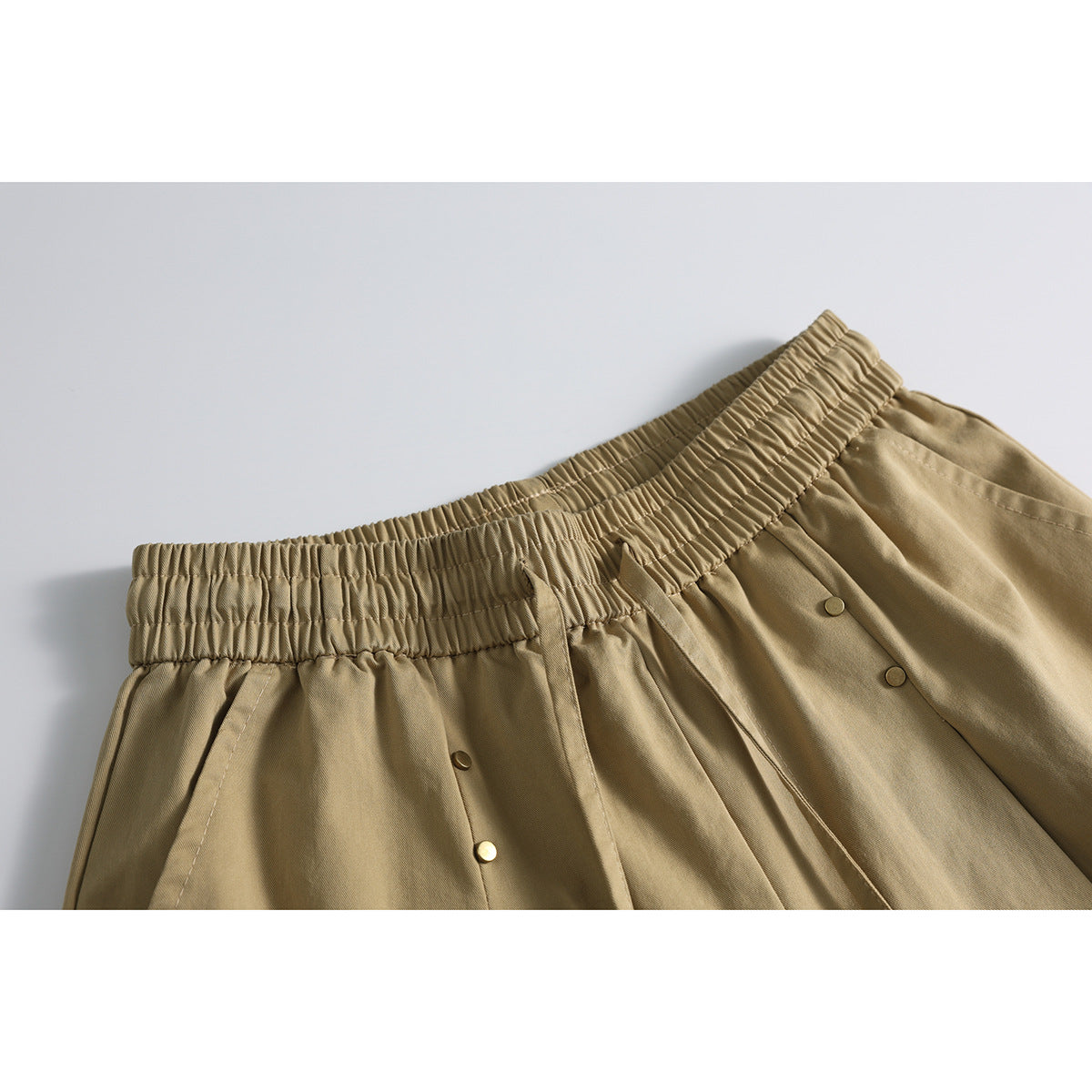 Close-up of tan shorts showing a gathered elastic waistband, drawstrings, pleats and small metal buttons.