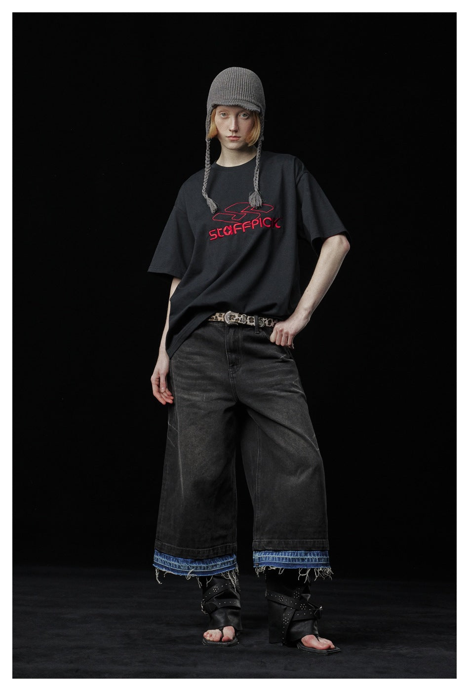 Full-length model wearing a black T-shirt with red chest lettering, wide dark jeans, and a gray knit cap.