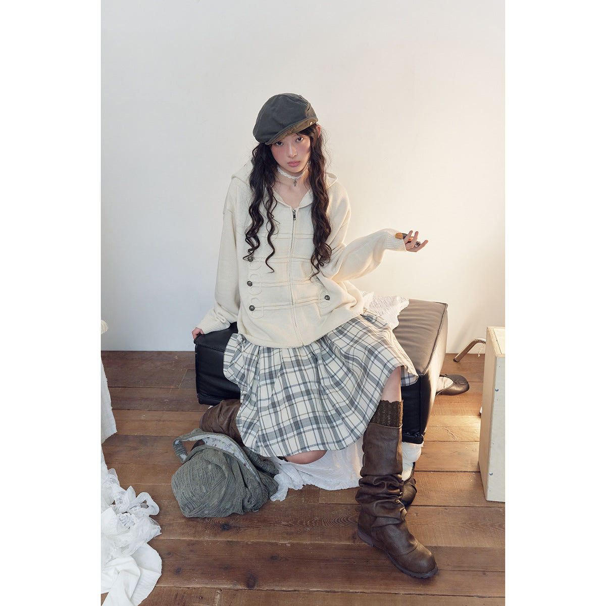 Seated model wearing a cream hooded zip sweater with decorative loop-and-button detailing and a gray plaid skirt.