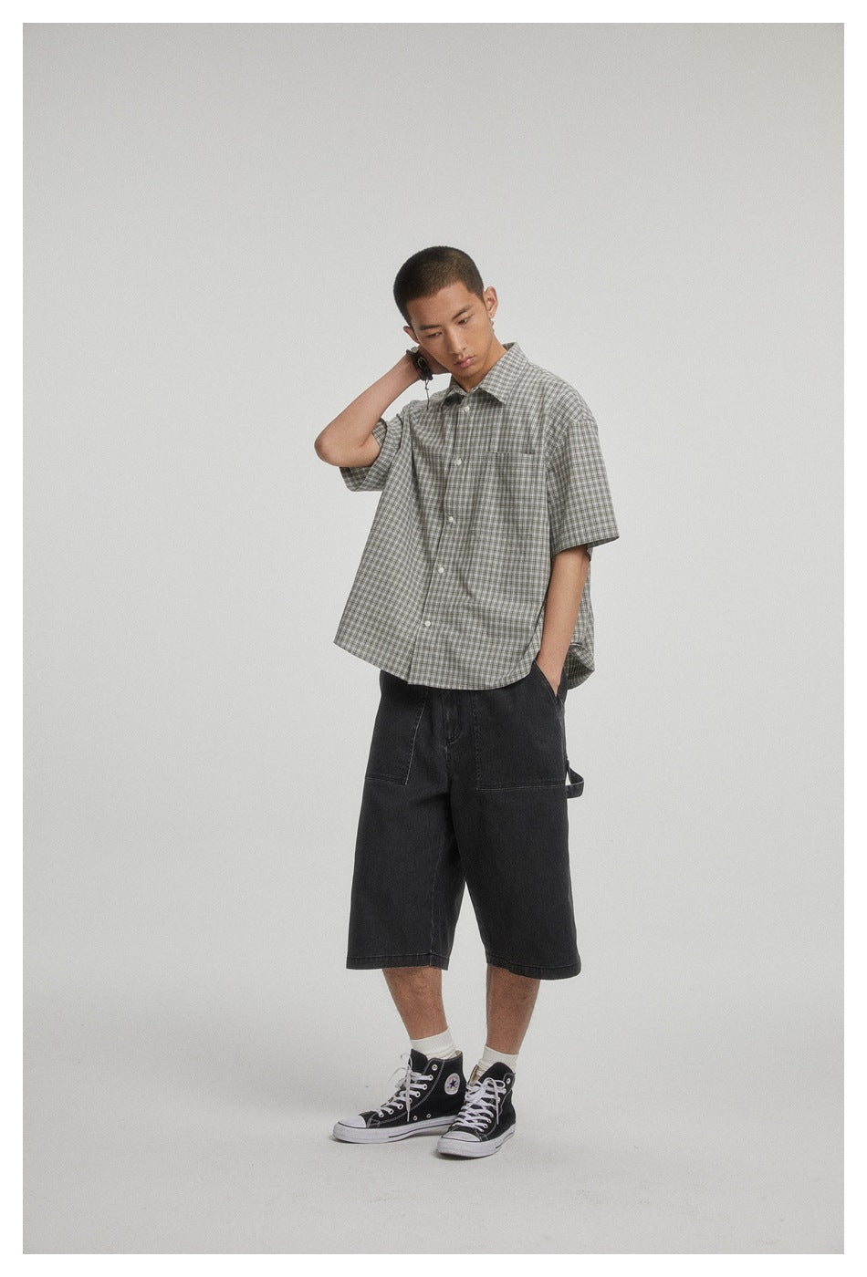 Model wearing a relaxed gray plaid short-sleeve button-up shirt with a chest pocket, posed with one hand behind his neck.