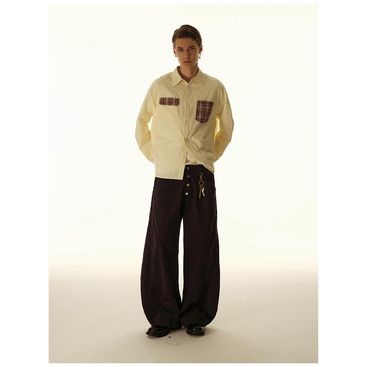 Full-length model wearing a cream button-up shirt with plaid chest pockets and wide dark trousers.