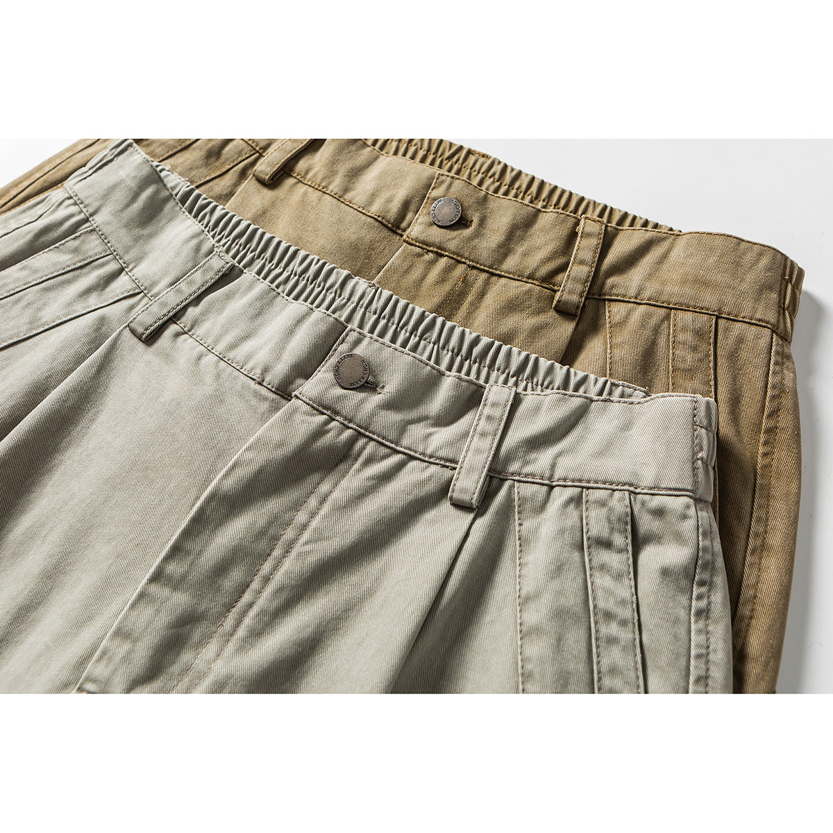 Close-up of two pairs of pleated trousers in light gray and tan, showing elastic waistbands, belt loops, and buttons.