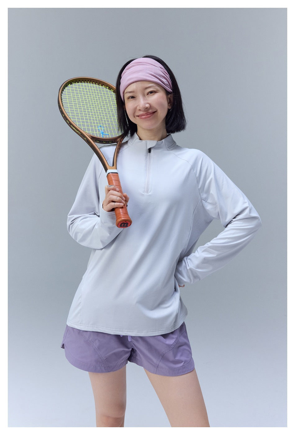 Model wearing a pale gray quarter-zip top and lavender shorts, posing with a tennis racket.