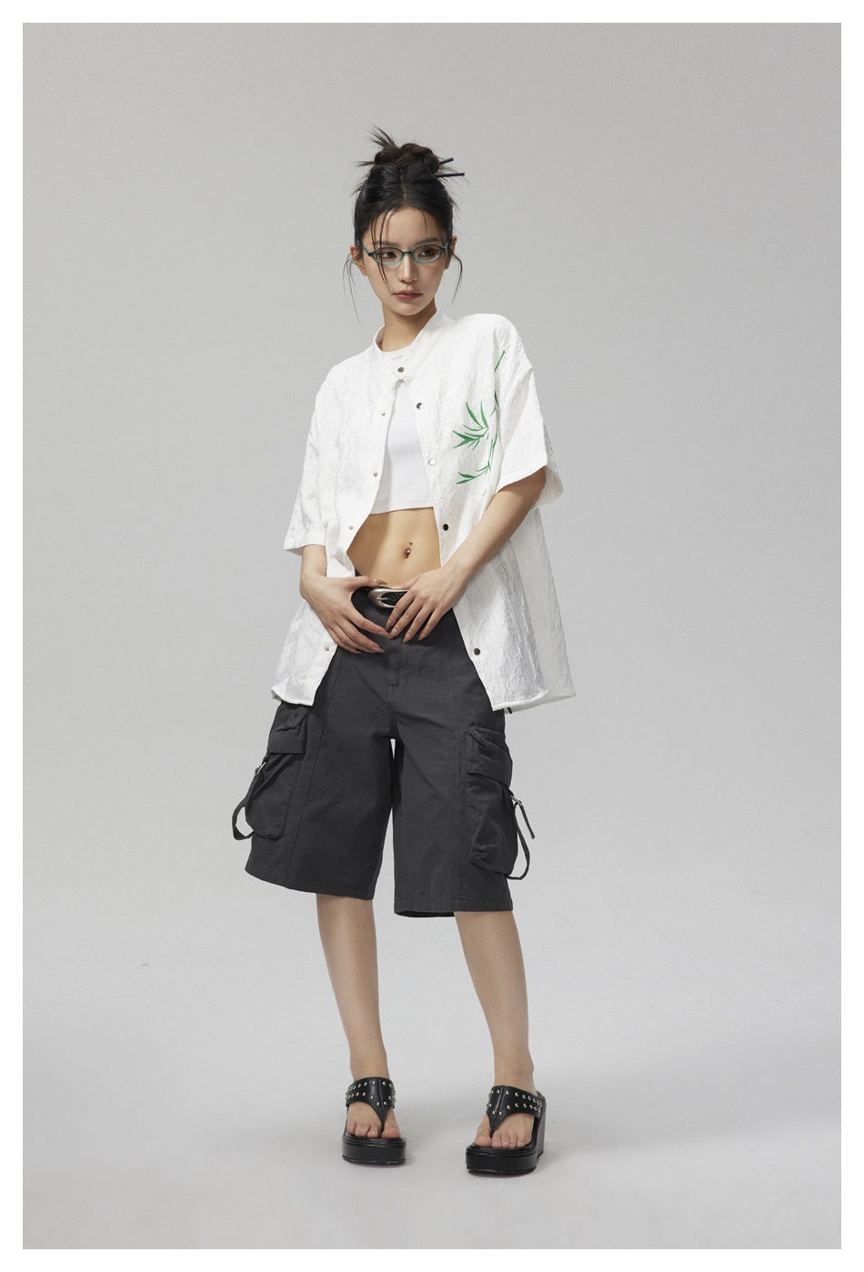 Model wearing the white bamboo-embroidered short-sleeve shirt open over a white crop top with gray cargo shorts