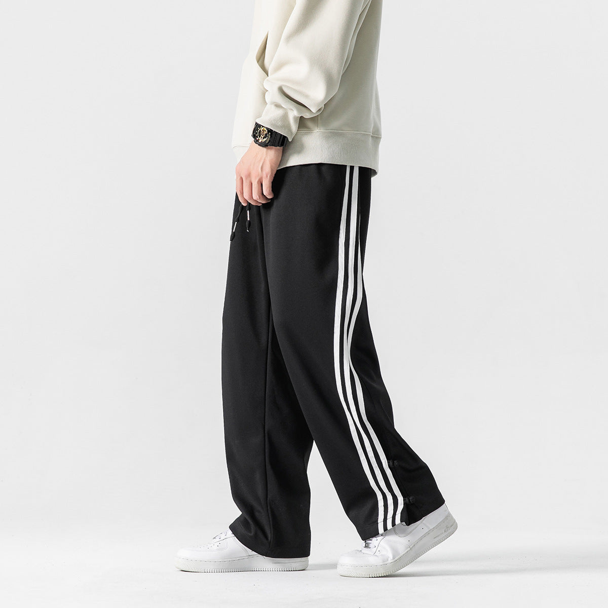 Cropped model wearing loose black track pants with three white side stripes and snap details near the hems.