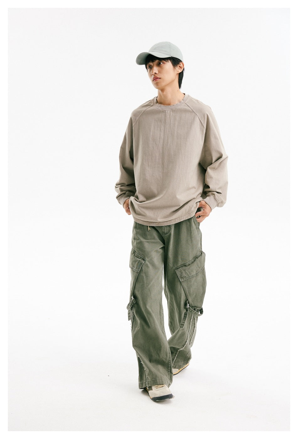 Model wearing a loose taupe crewneck and olive cargo pants, walking toward the camera in a light cap.