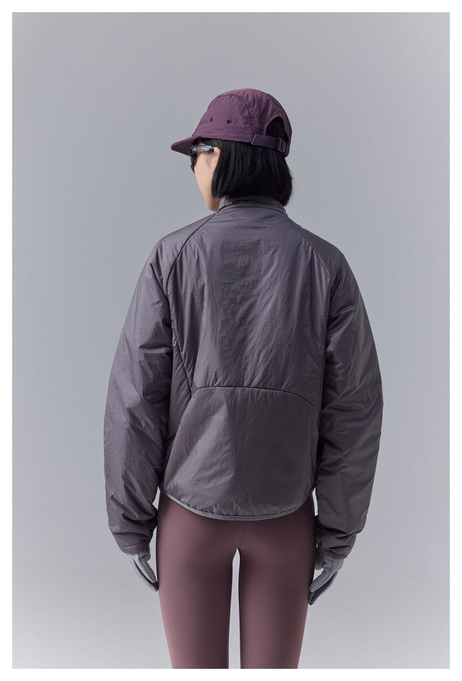 Rear view of a model wearing a gray-plum jacket and mauve leggings, showing the jacket's curved hem and back panel.