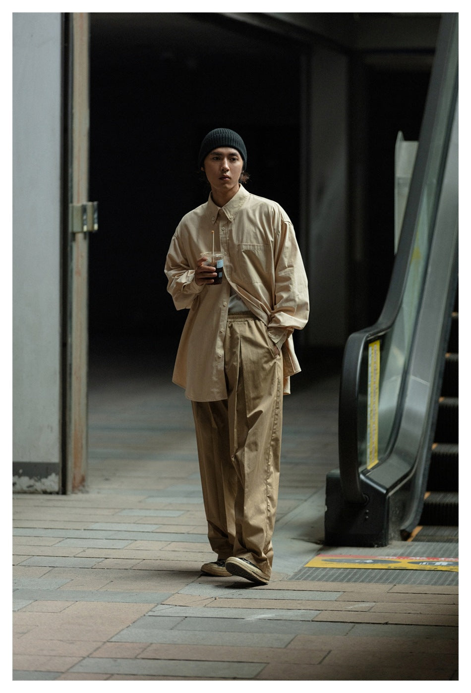 Front view of a model wearing a loose beige button-up shirt and tan trousers with a black beanie.